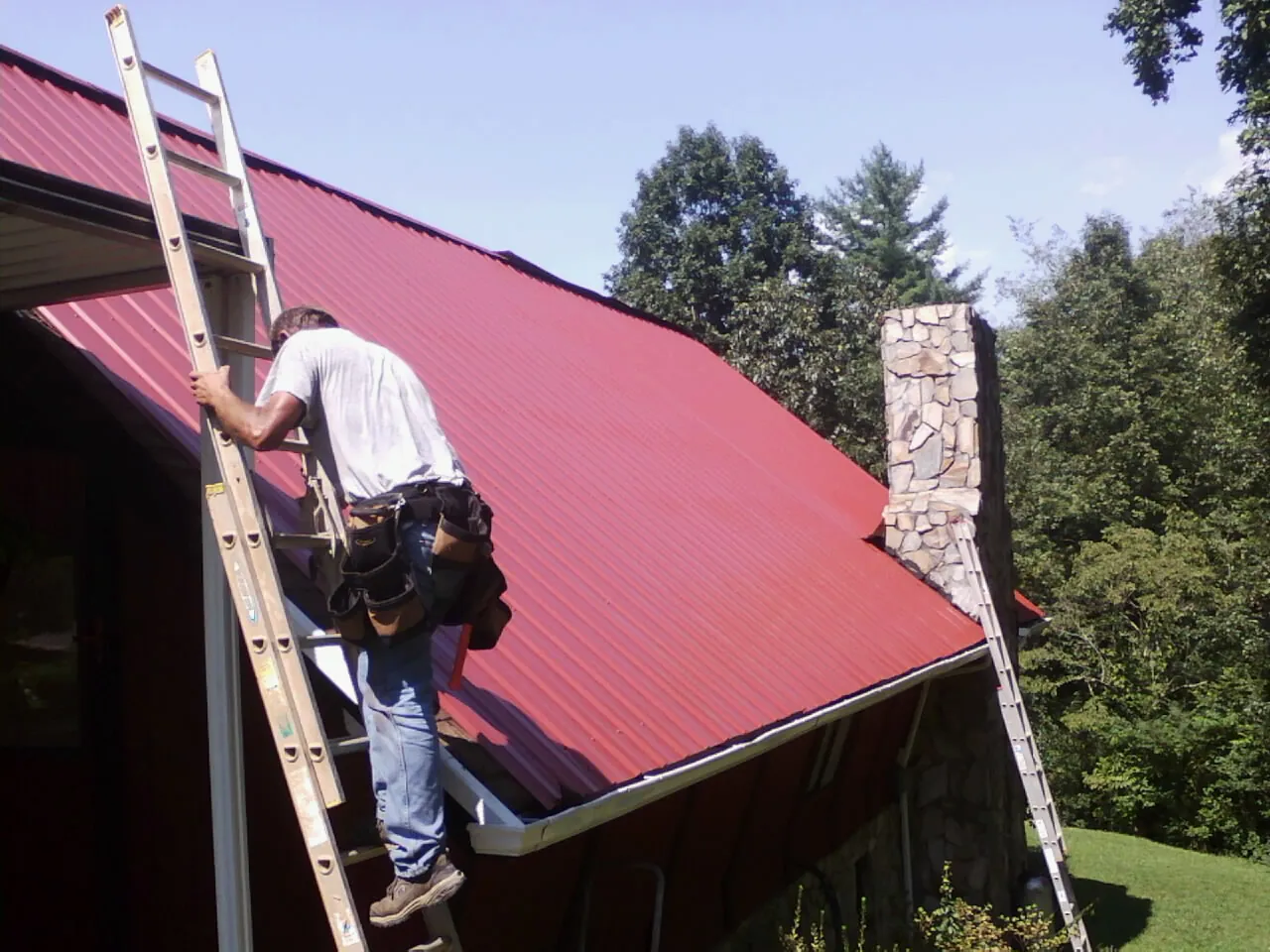 Roofer on a ladder beside a red metal roof and stone chimney — Soffit & Fascia Repair by Roofing Brothers in Sausalito